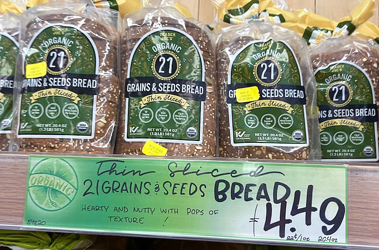Display of organic 21 grains and seeds bread packages in a store, with a price tag of $4.49 and a sign describing its texture as hearty and nutty.