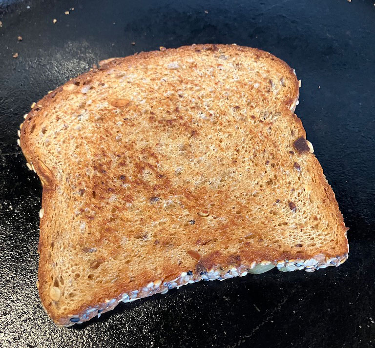A slice of toasted whole grain bread with a golden-brown crust placed on a black skillet.