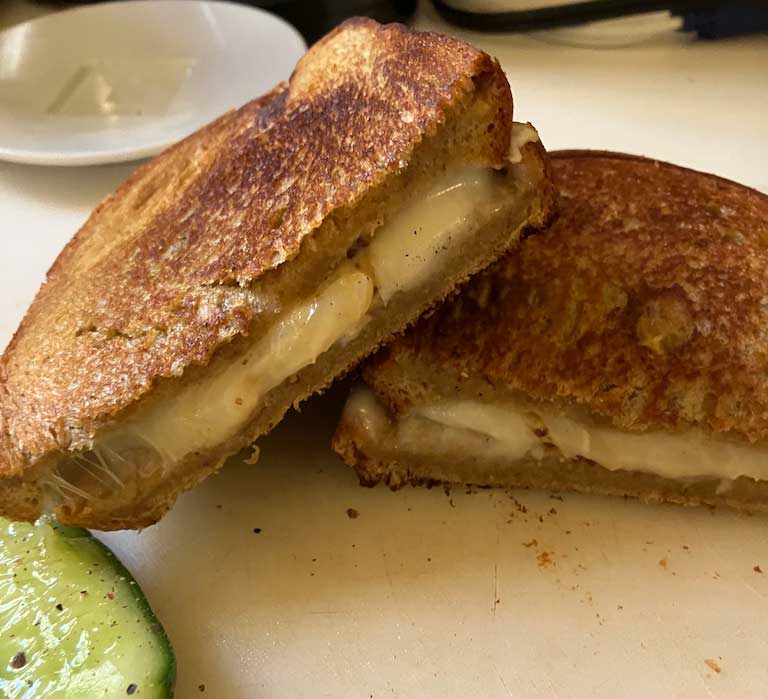 Close-up of a grilled cheese sandwich, cut in half, revealing melted cheese between two slices of toasted bread, with a slice of cucumber on the side.